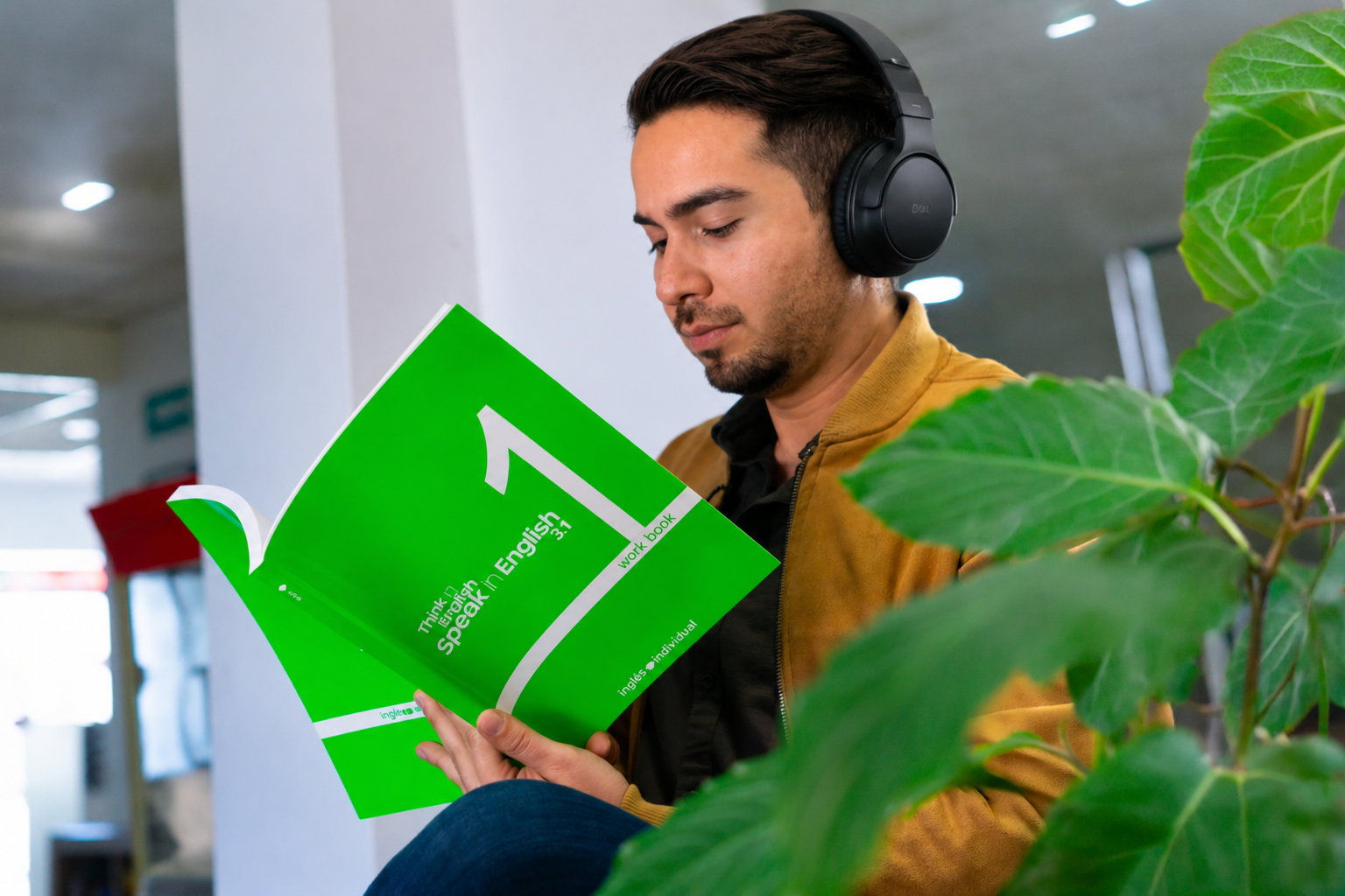 Man with headphones reading a green workbook titled Speak in English 3.1 indoors near a plant.