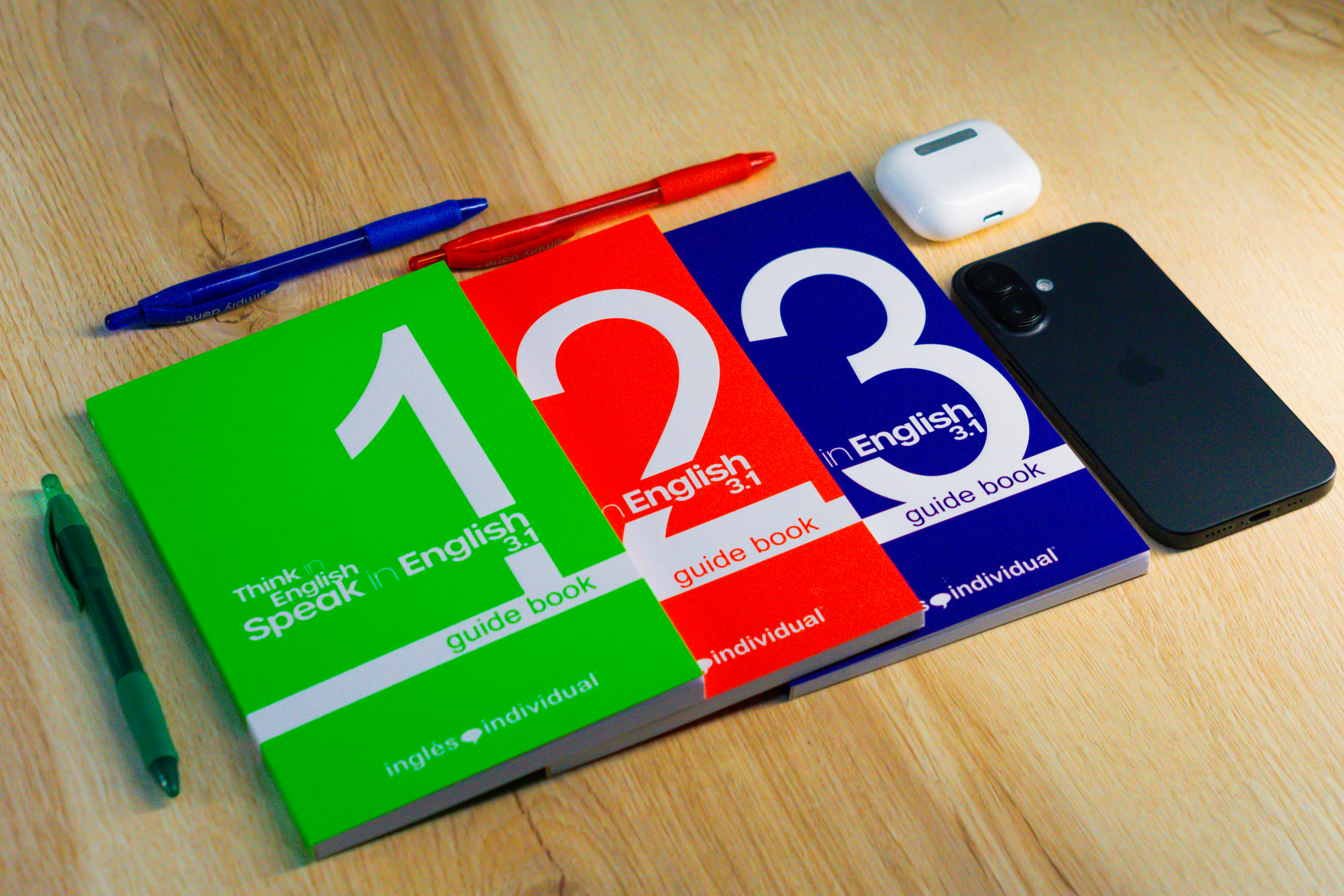 Three colorful English guide books numbered 1, 2, and 3 next to pens, wireless earbuds, and a black smartphone.