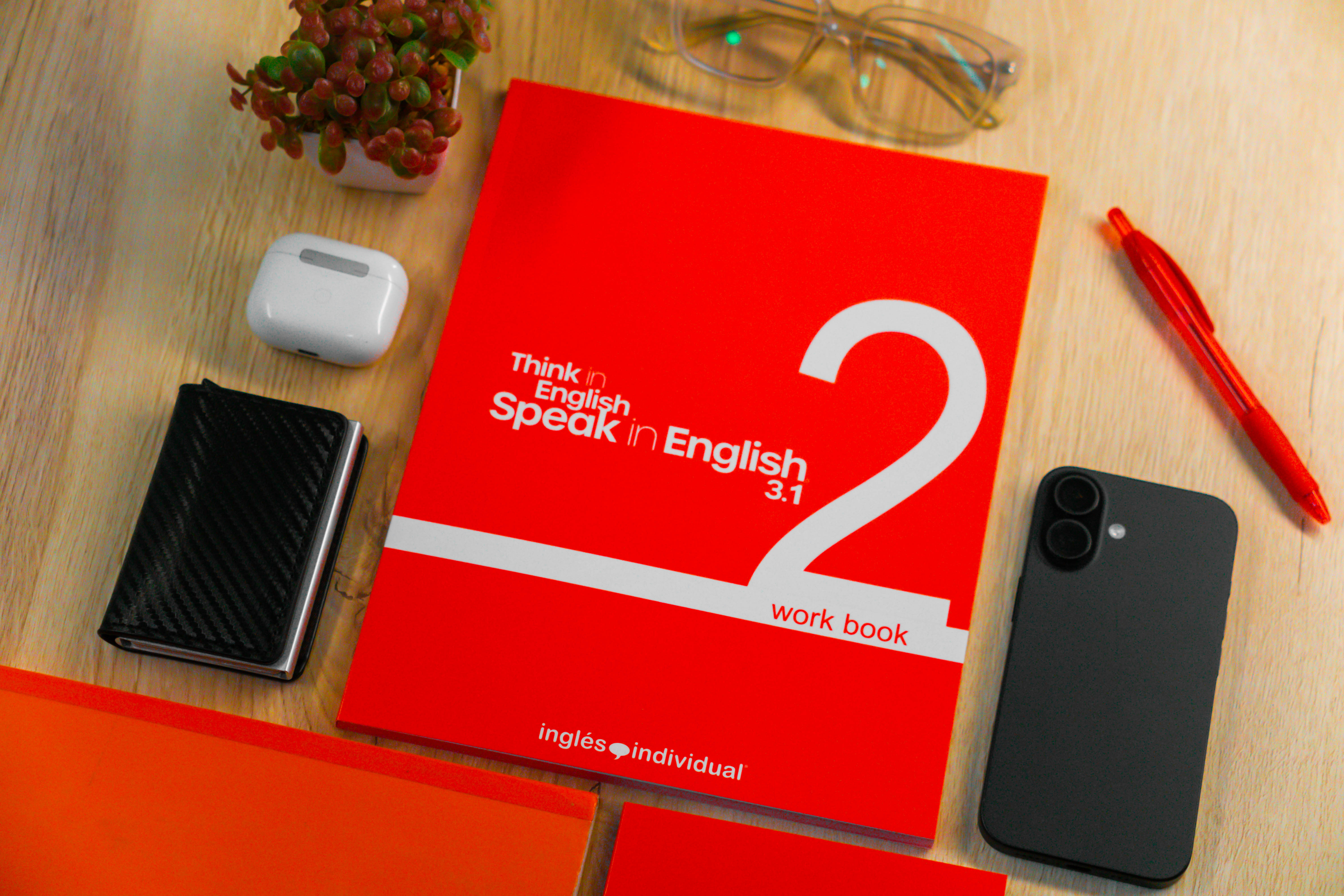 Red workbook titled 'Think in English, speak in English 3.1 work book' on a wooden desk with tech items.