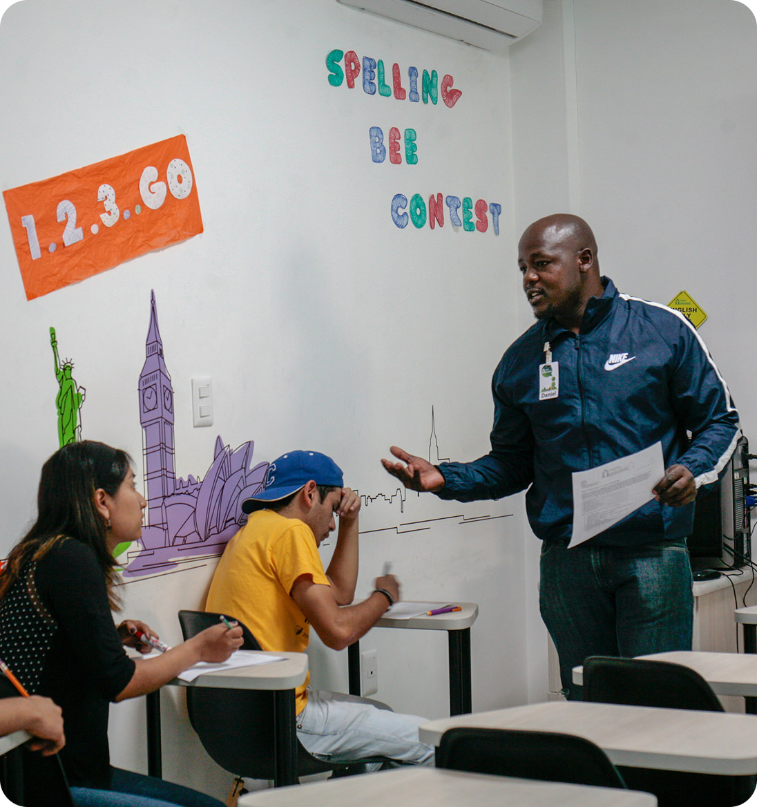 Teacher in navy jacket talking to two students seated in a classroom during a spelling bee contest.